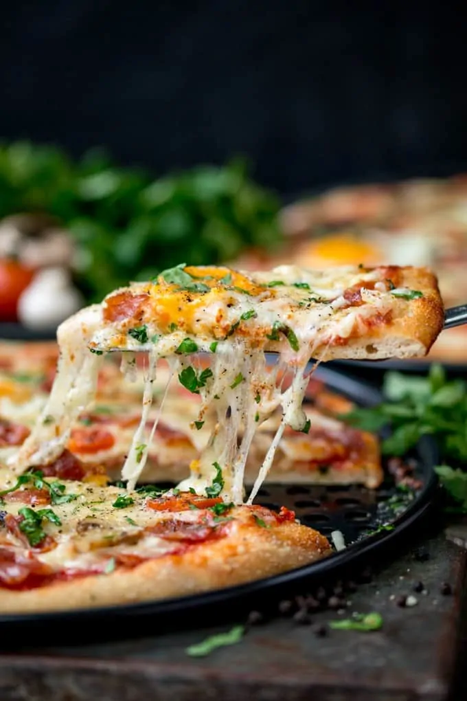 Side-on image of egg-topped breakfast pizza slice being lifted from the pizza. Melted cheese is stretching between the pizza and pizza slice.