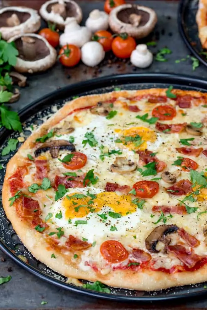 Slightly cut off image of breakfast pizza with eggs, bacon, tomatoes and mushrooms. Uncut mushrooms, tomatoes and parsley in the background.