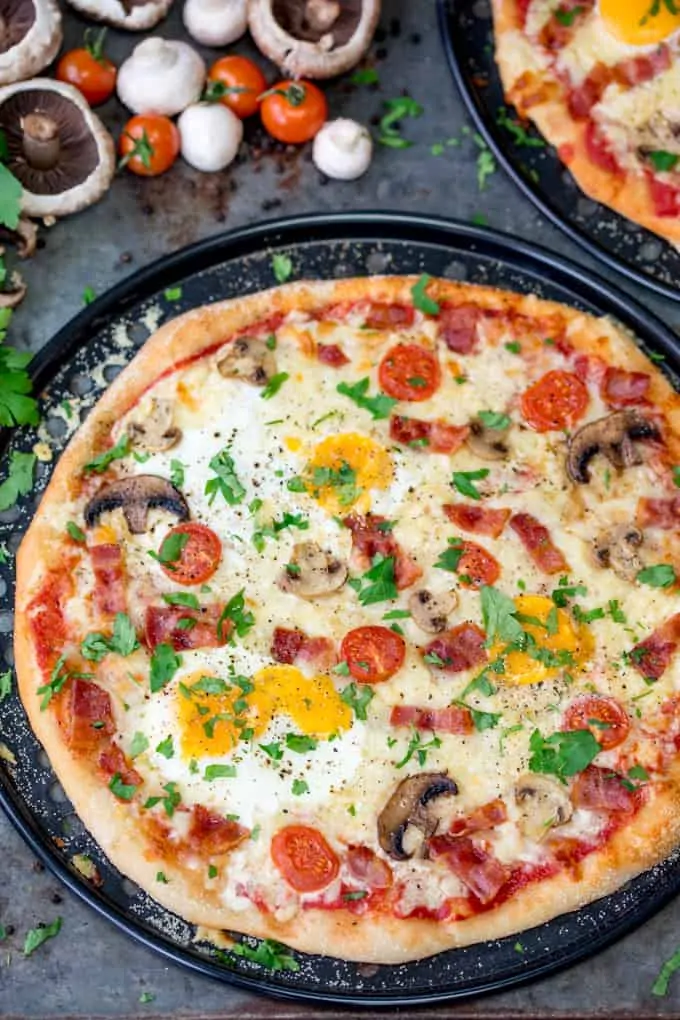 Slightly cut off image of breakfast pizza with eggs, bacon, tomatoes and mushrooms. Uncut mushrooms, tomatoes and parsley in the background.