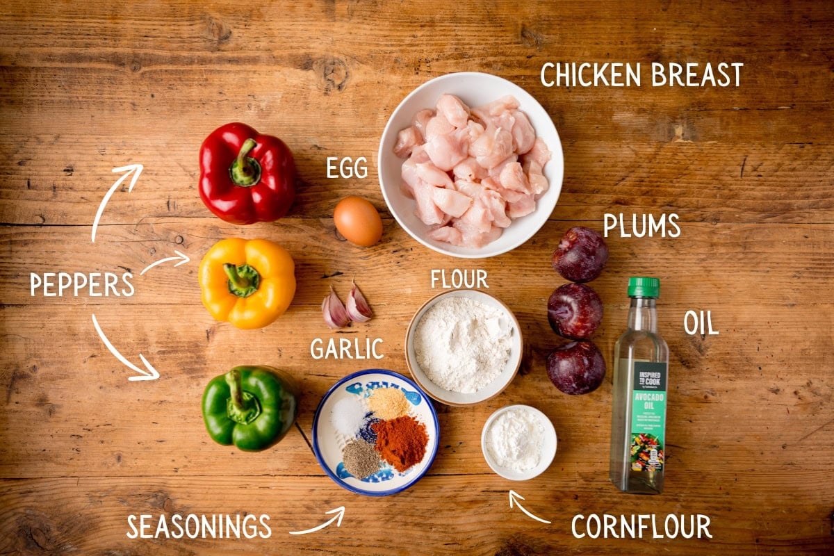 Ingredients on a wooden table for making Crispy Chicken plum stir fry, including red, yellow, and green peppers, plums, chicken pieces, an egg, flour, garlic, spices, cornflour, and oil. Each ingredient is labelled.