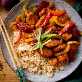 Chinese plum chicken stir fry and rice in a shallow bowl with strips of spring onion on top. There is a set of chopsticks on the side of the bowl. The bowl is on a wooden table.
