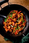 Overhead image of Chinese plum chicken stir fry with sliced peppers in a dark wok. A green napkin is just in shot on the handle of the wok.