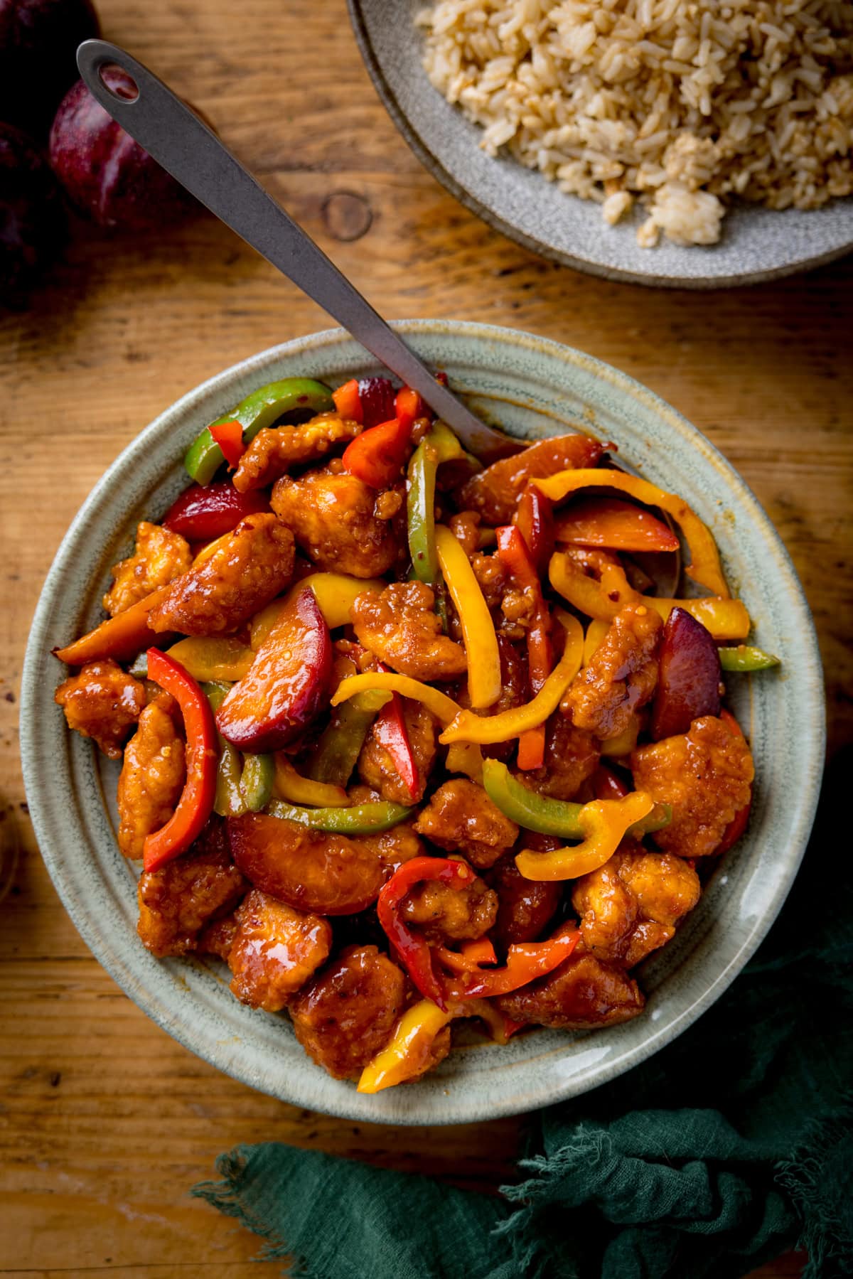 A bowl of Chinese chicken plum stir-fry with red, yellow, and green peppers in a bowl on a wooden table. A green napkin is at the bottom of the shot.