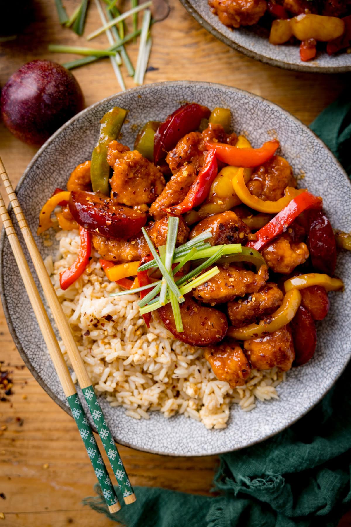 Chinese plum chicken stir fry and rice in a shallow bowl with strips of spring onion on top. There is a set of chopsticks on the side of the bowl. The bowl is on a wooden table next to a green napkin.