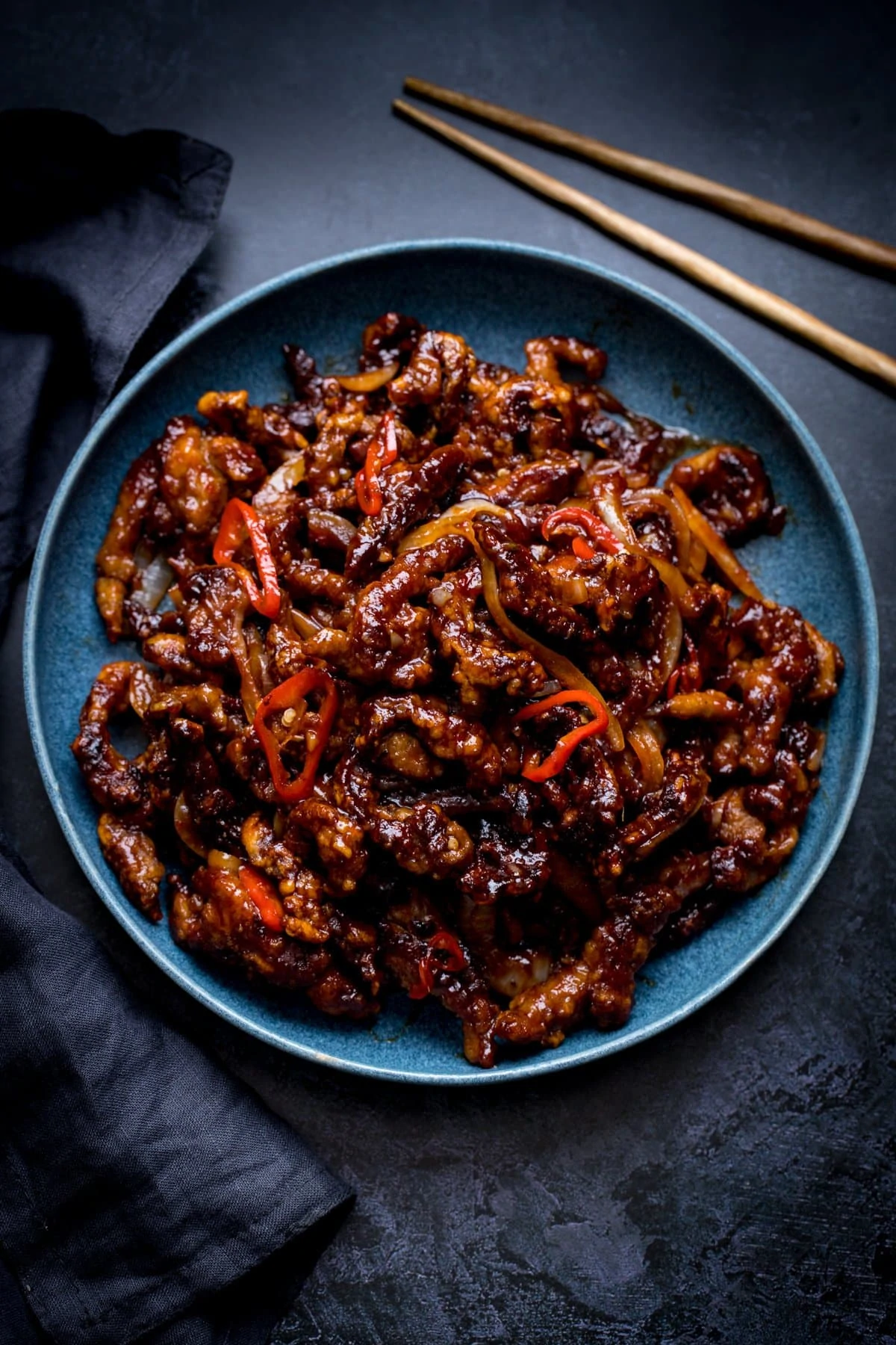 crispy chilli beef in a blue bowl on a dark background. Dark napkin and set of chopsticks also in shot.