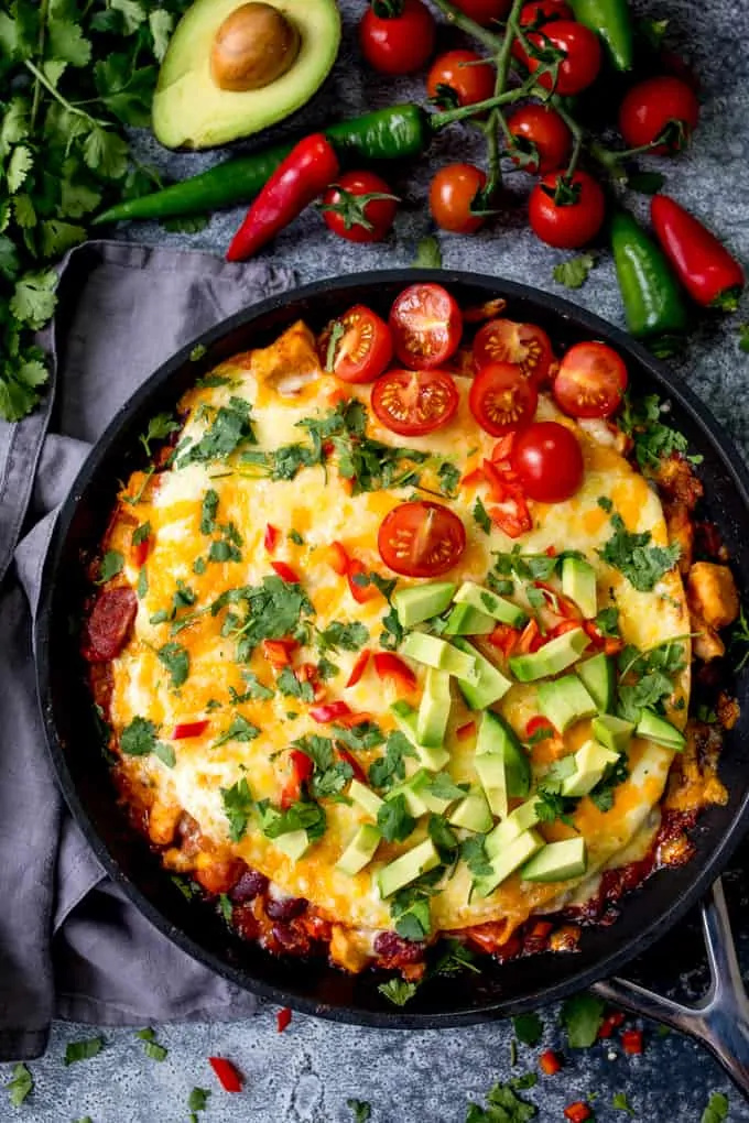 Overhead photo of chicken enchilada lasagna in a pan, topped with avocado, tomatoes and chopped chillies.
