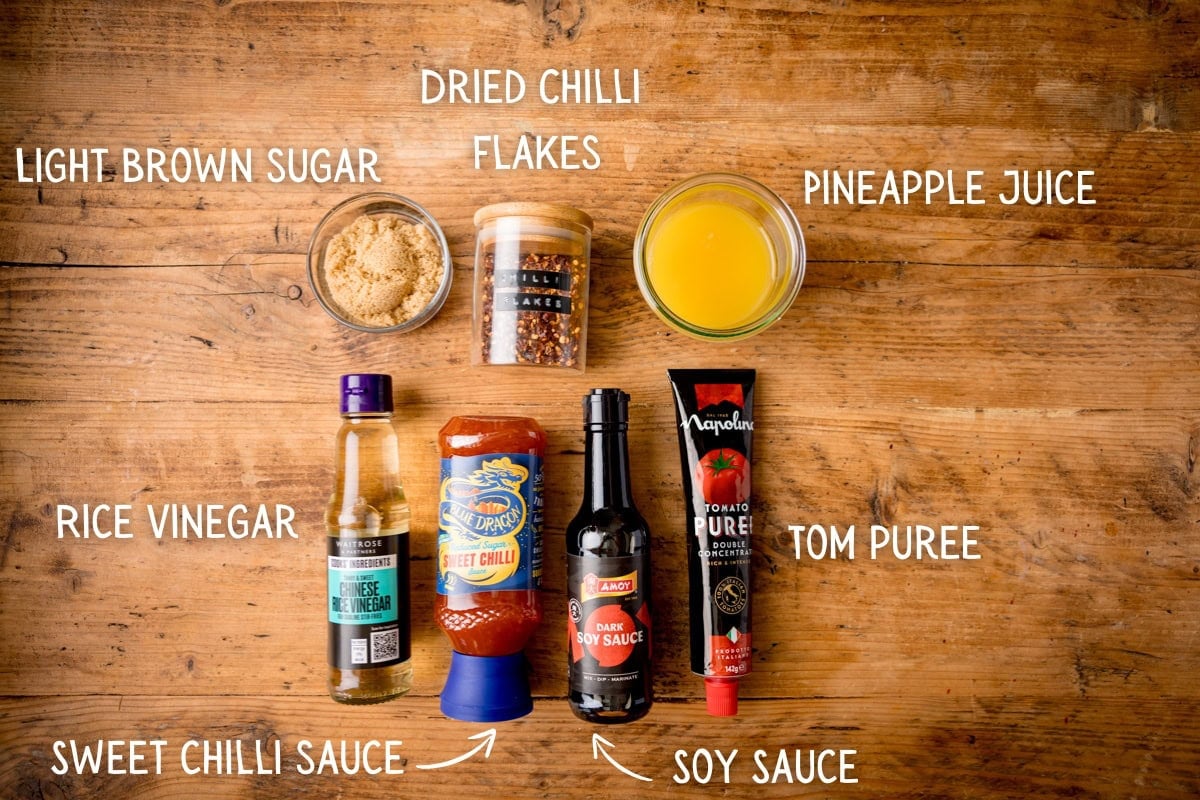 Ingredients for Chinese Plum Chicken Sauce, including sugar, chilli flakes, pineapple juice, rice vinegar, sweet chilli sauce, soy sauce, and tomato purée, on a wooden surface. Each ingredient is labelled.