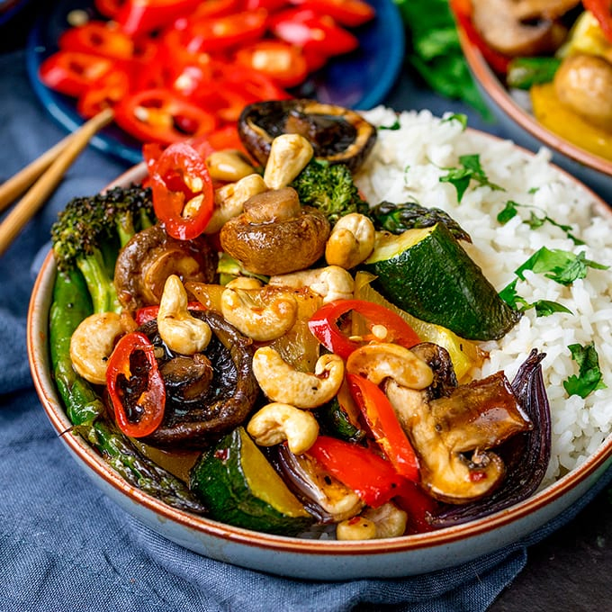 Bowl of vegetables in asian-style sauce with rice, cashew nuts and chopped chillies.