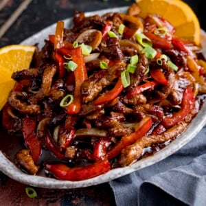 Crispy orange beef with peppers and onions on a flat oval plate.  There are orange slices on the plate.