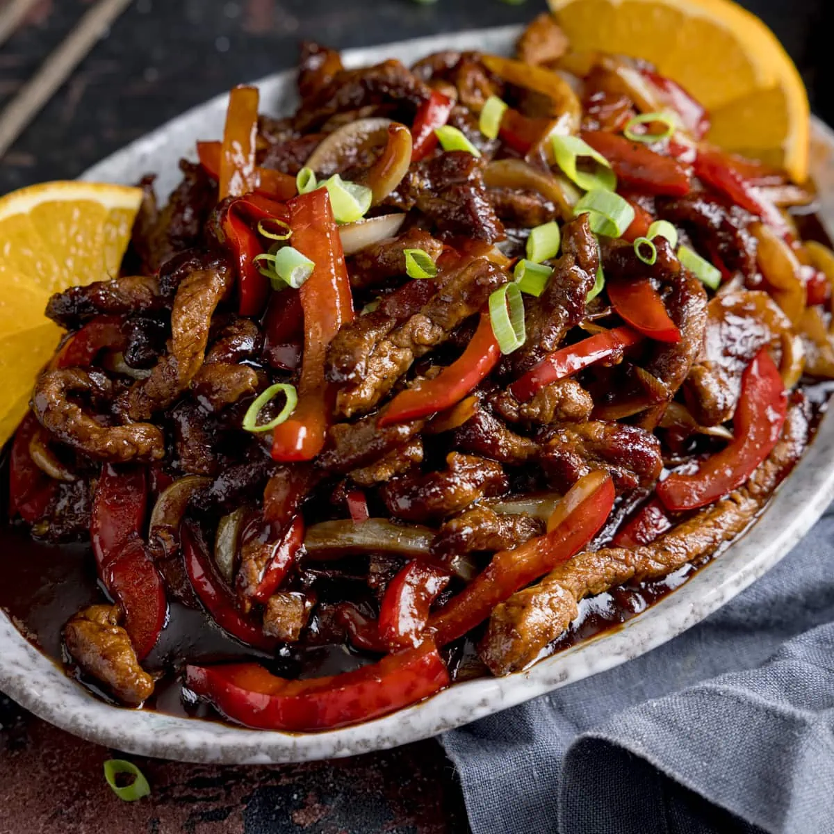 Crispy orange beef with peppers and onions on a shallow oval plate. There are slices of orange on the plate.