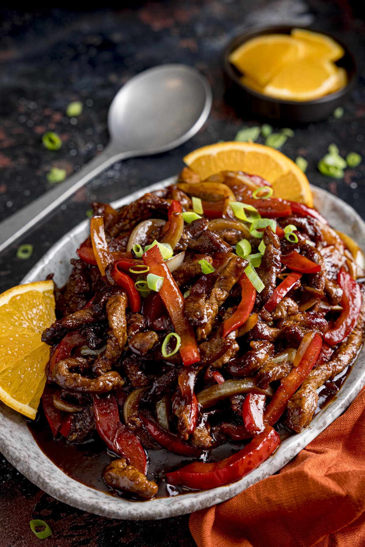 Crispy orange beef with peppers and onions on a shallow oval plate. There are slices of orange and spring onions scattered around and a spoon in the background.