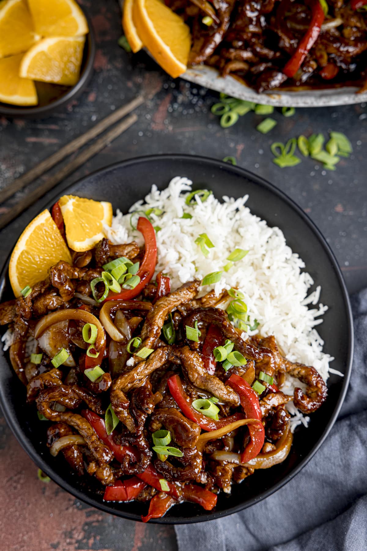 Overhead of crispy orange beef with peppers and onions on a dark plate with boiled rice. There are slices of orange and spring onions scattered around and a pair of chopsticks next to the plate.