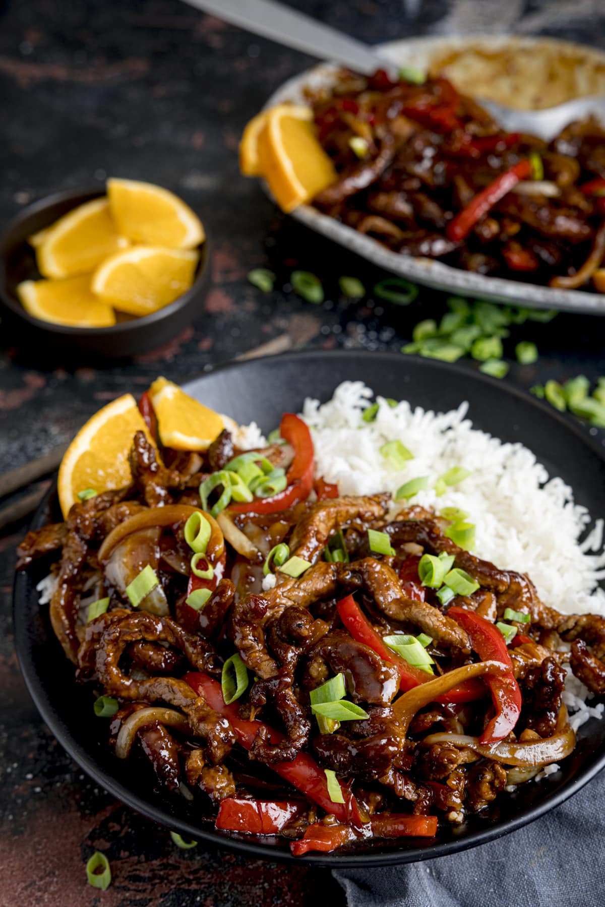 Crispy orange beef with peppers and onions on a dark plate with boiled rice.  Orange slices and spring onions are scattered, and the serving platter can be seen in the background.