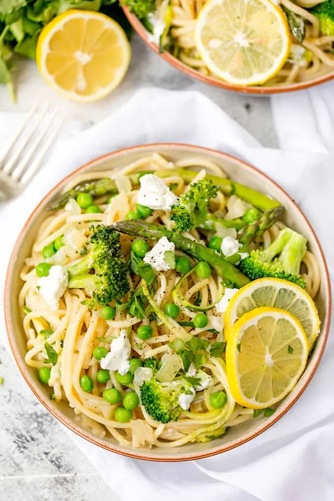 Bowl of spaghetti with spring vegetables and goats cheese with lemon slices, on a white background