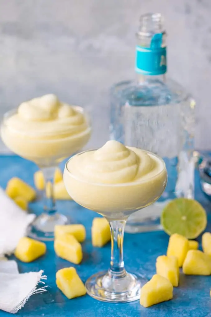 Two glasses of dole whip margarita on blue background. Pineapple chunks, half a lime and bottle of tequila in background.