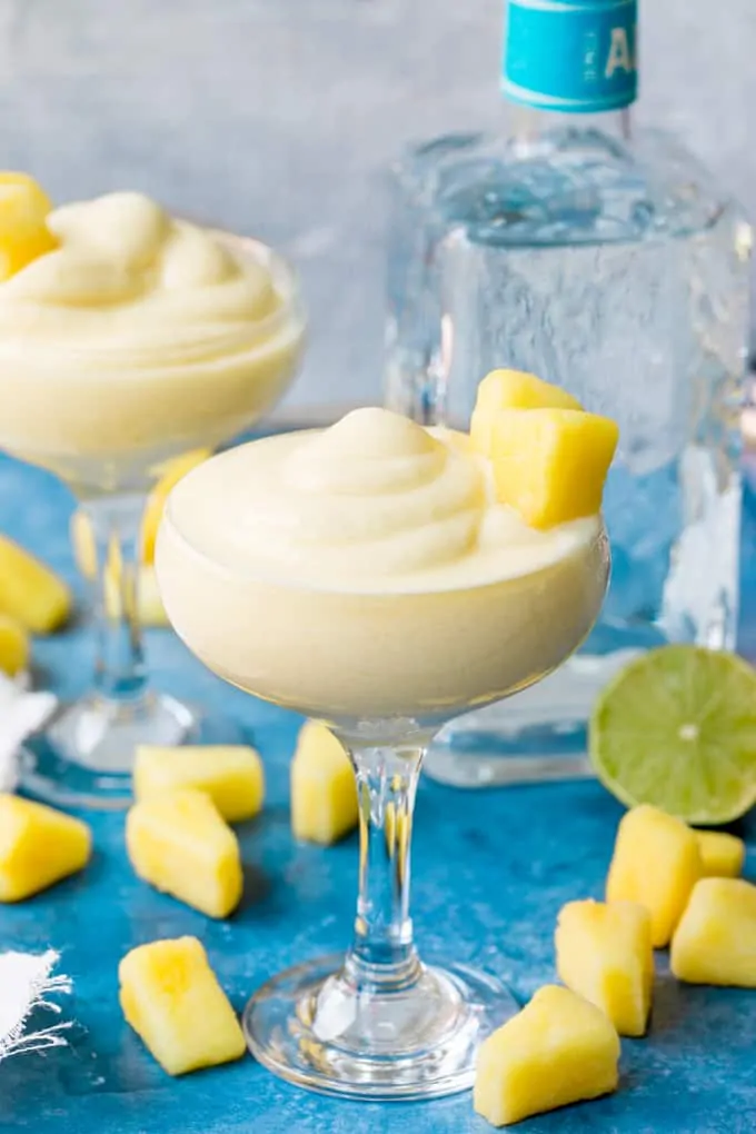 Two glasses of dole whip margarita with pineapple chunk decoration, on blue background. Pineapple chunks, half a lime and bottle of tequila in background.