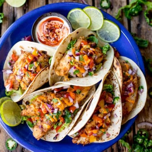 Overhead square image of 6 crispy fish tacos on a blue plate, topped with pico de gallo and sriracha mayo.