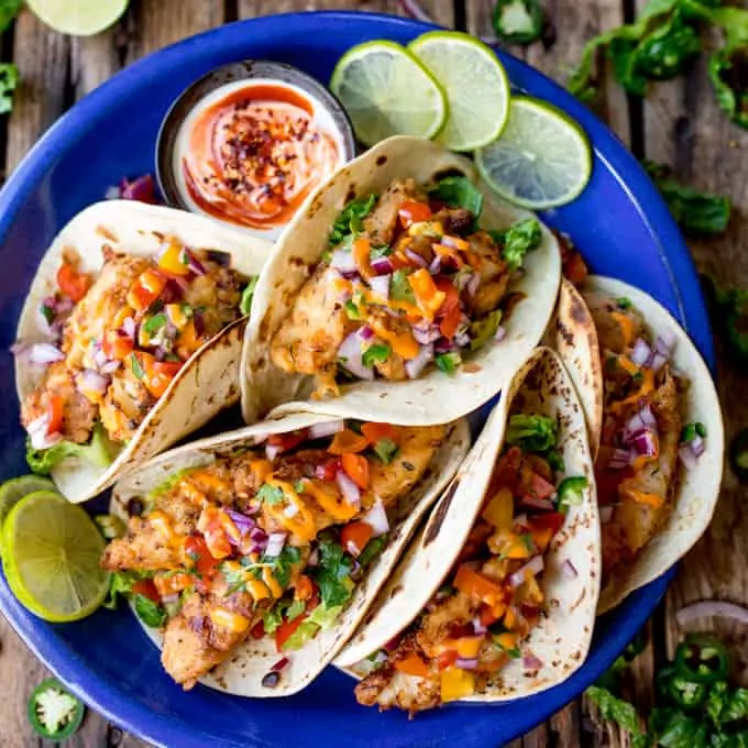 Overhead square image of 6 crispy fish tacos on a blue plate, topped with pico de gallo and sriracha mayo.