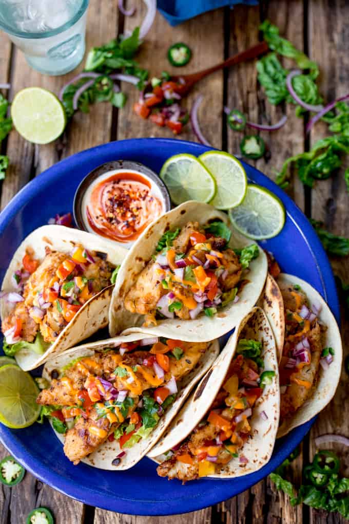 Tall image of 6 crispy fish tacos on a blue plate on a wooden background. Tacos are topped with pico de gallo and sriracha mayo. Further ingredients in background.