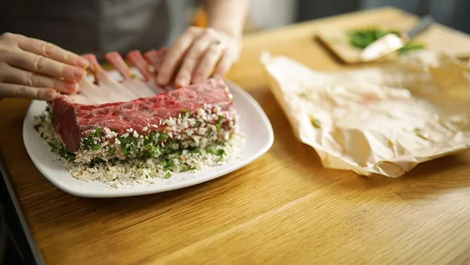 Rack of lamb being rolled in a herbed breadcrumb coating on a white plate on a wooden surface.