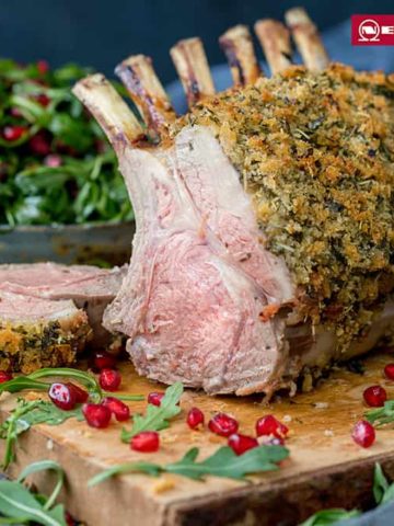 Herb crusted rack of lamb on a wooden board with a slice taken off - showing pink centre. Rocket and pomegranate seeds strewn around the board.