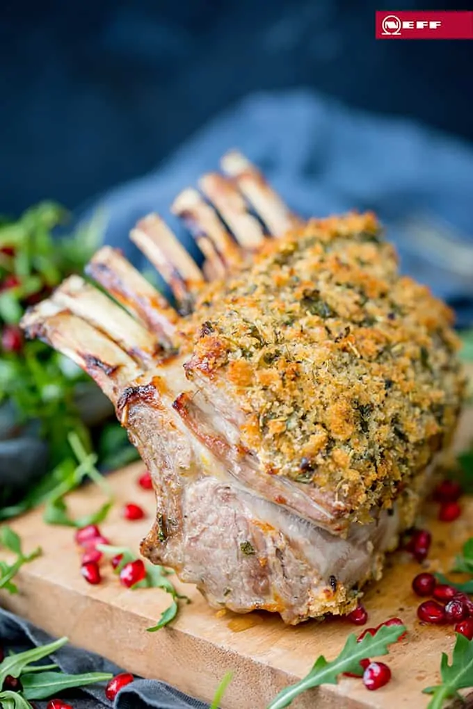 Tall image of a herb crusted rack of lamb on a wooden board. Rocket and pomegranate seeds strewn around the board.