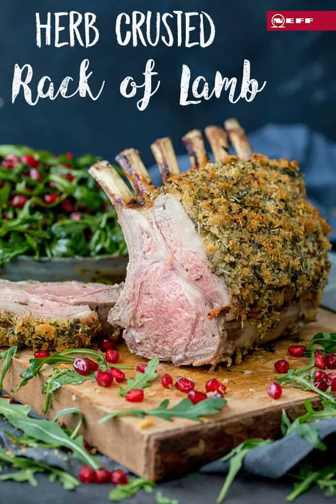 Tall image of a herb crusted rack of lamb on a wooden board with a slice taken off - showing pink centre. Rocket and pomegranate seeds strewn around the board.. Writing at the top of the image with the recipe title..