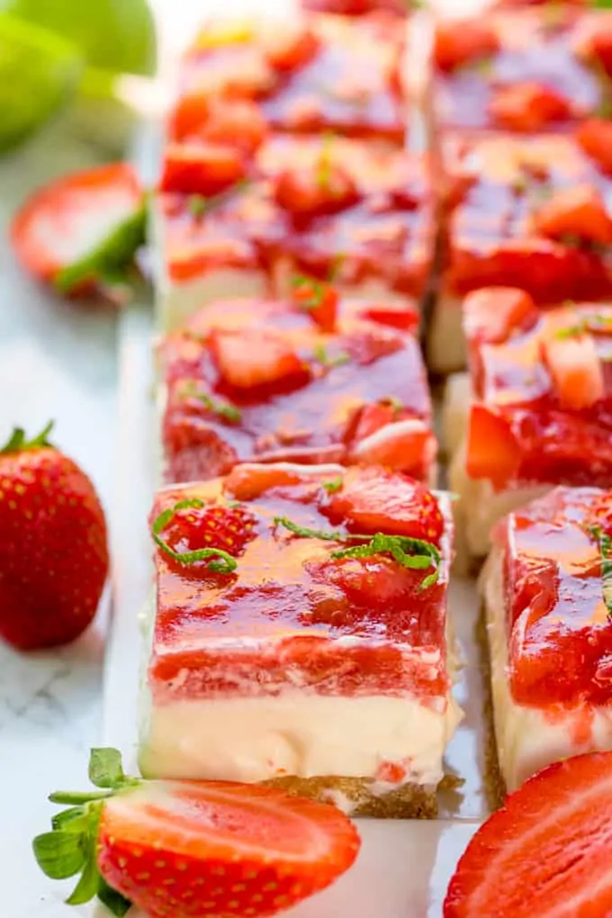 Tall image of strawberry and rhubarb cheesecake bars with jelly topping. Strawberries scattered around.