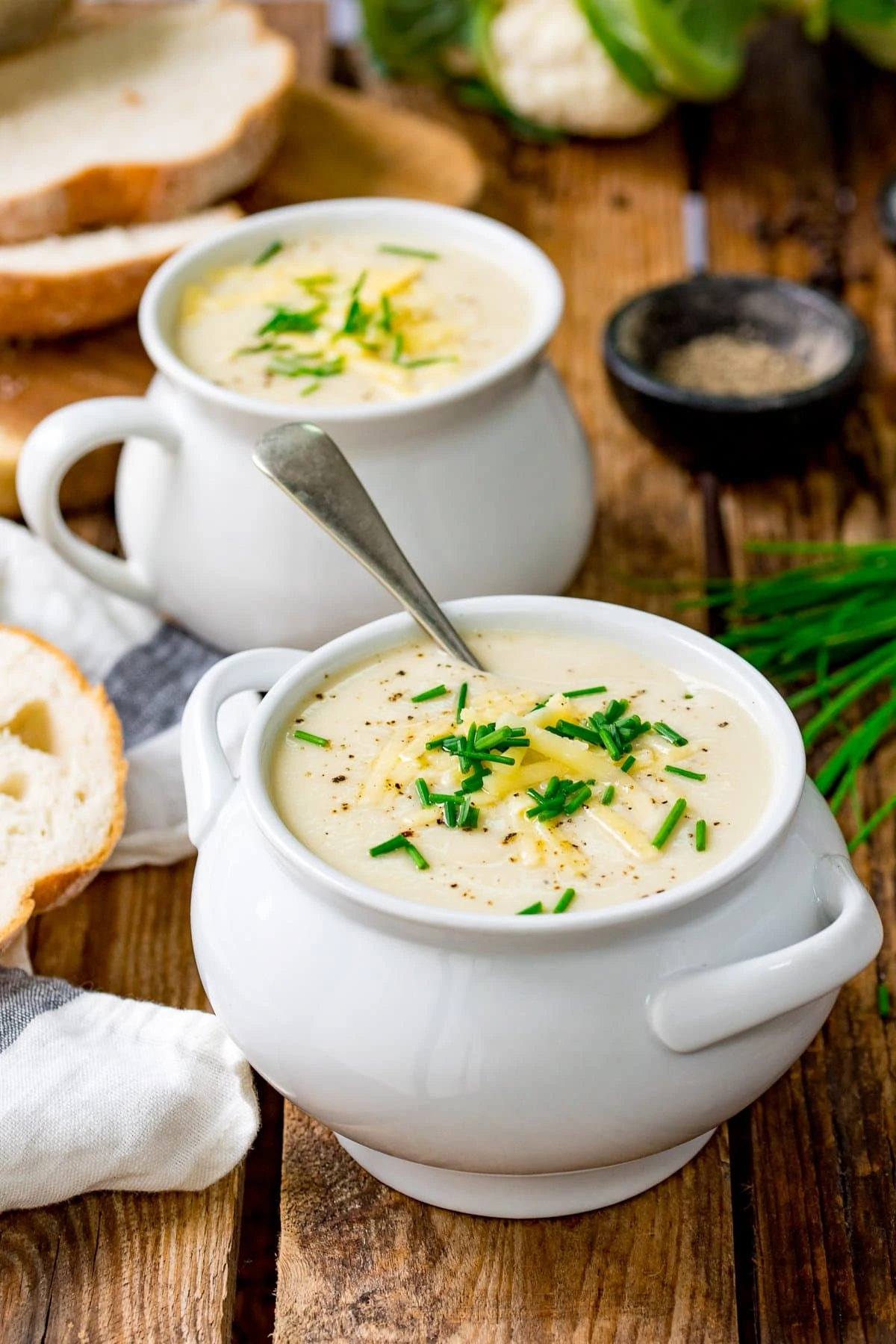 Two white bowls filled with cauliflower soup, topped with cheese and chives on a wooden table