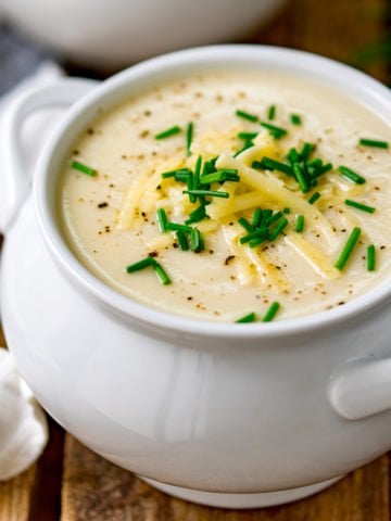 Close up of a white bowl filled with cauliflower soup and topped with chopped chives, grated cheese and black pepper