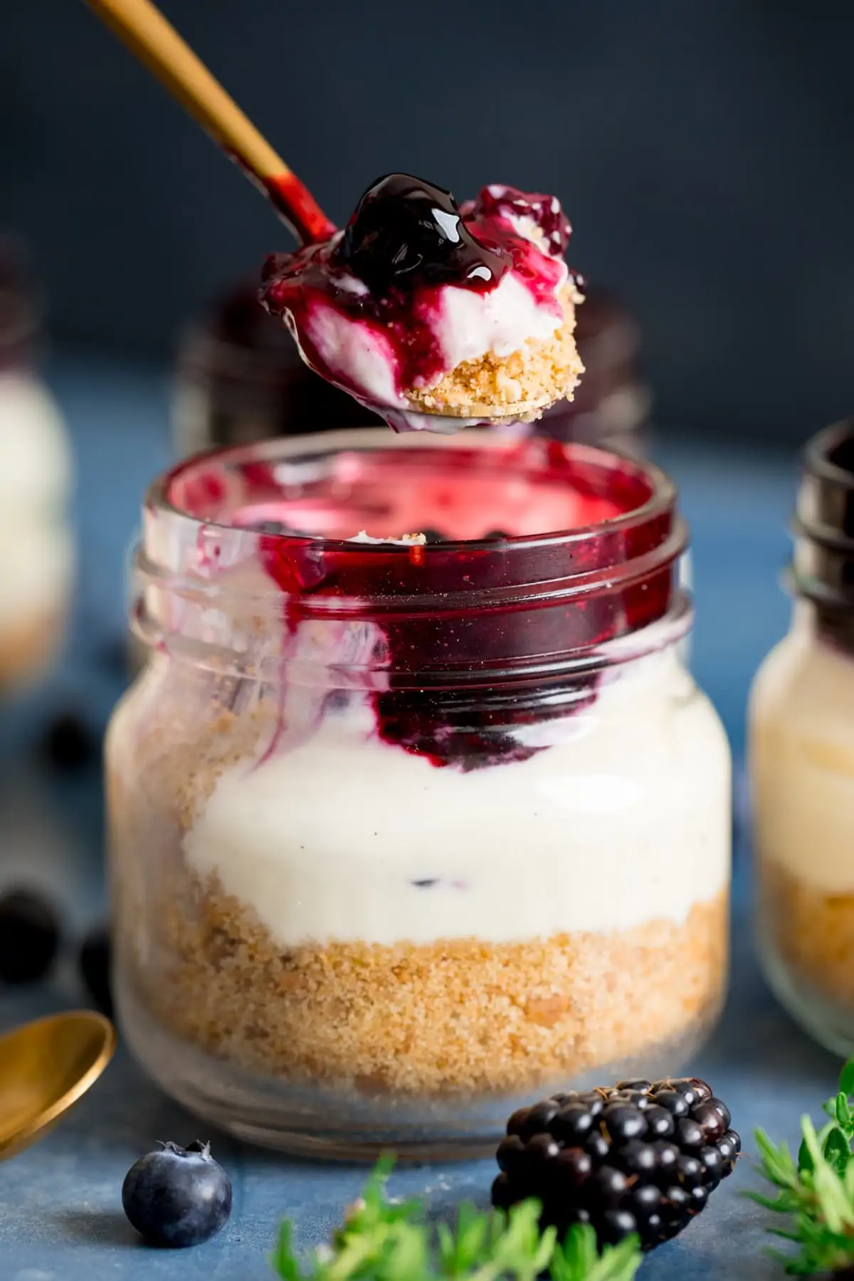 Spoonful being taken from a mason jar cheesecake on a blue background. Thyme leaves, blackberry and blueberries also in shot.