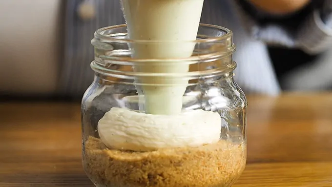 Cheesecake mixture being piped into a mason jar on top of a crushed biscuit base.