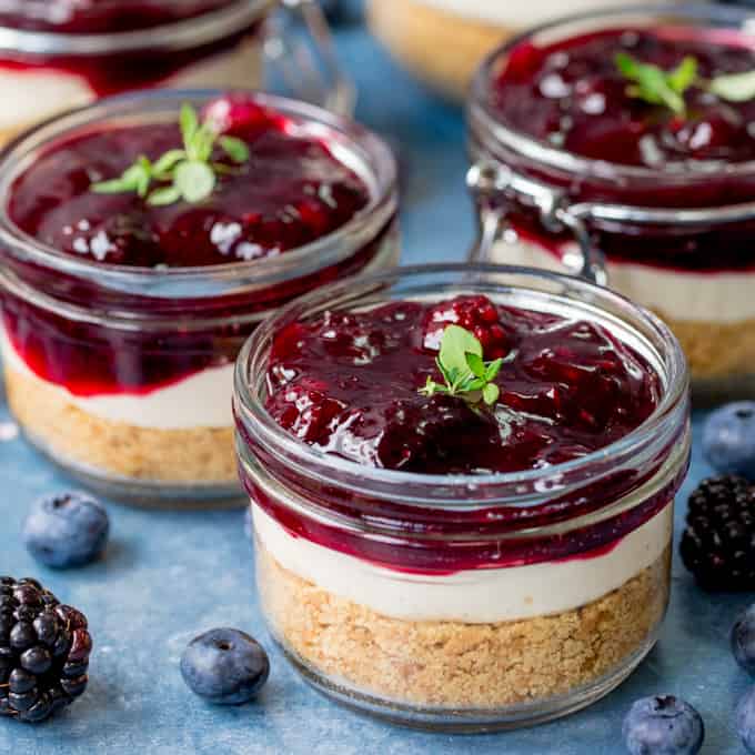 Square image of individual blueberry and blackberry cheesecakes in mason jars on blue background.