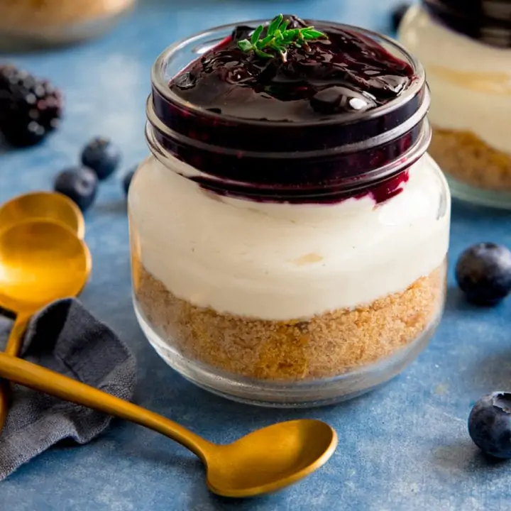 Individual berry cheesecake in a mason jar on a blue background next to a gold teaspoon