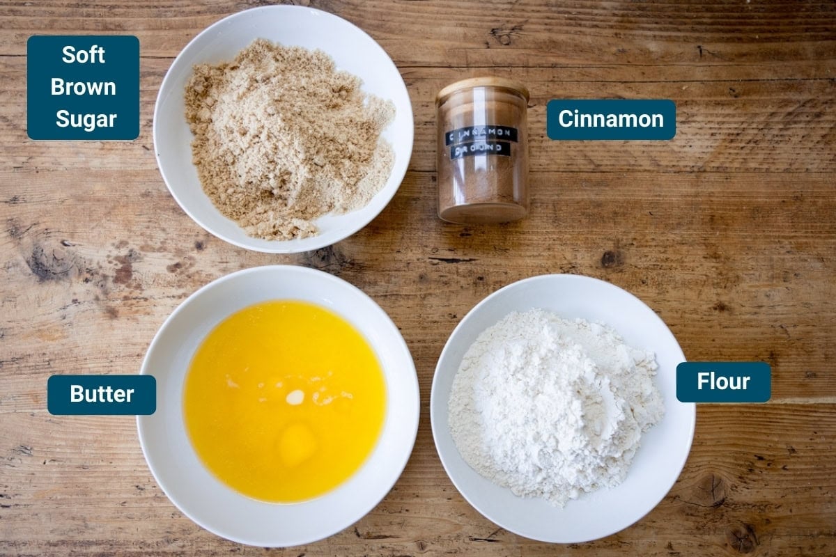 Four baking ingredients on a wooden table: soft brown sugar in a bowl, cinnamon in a jar, melted butter in a bowl, and flour in a bowl.