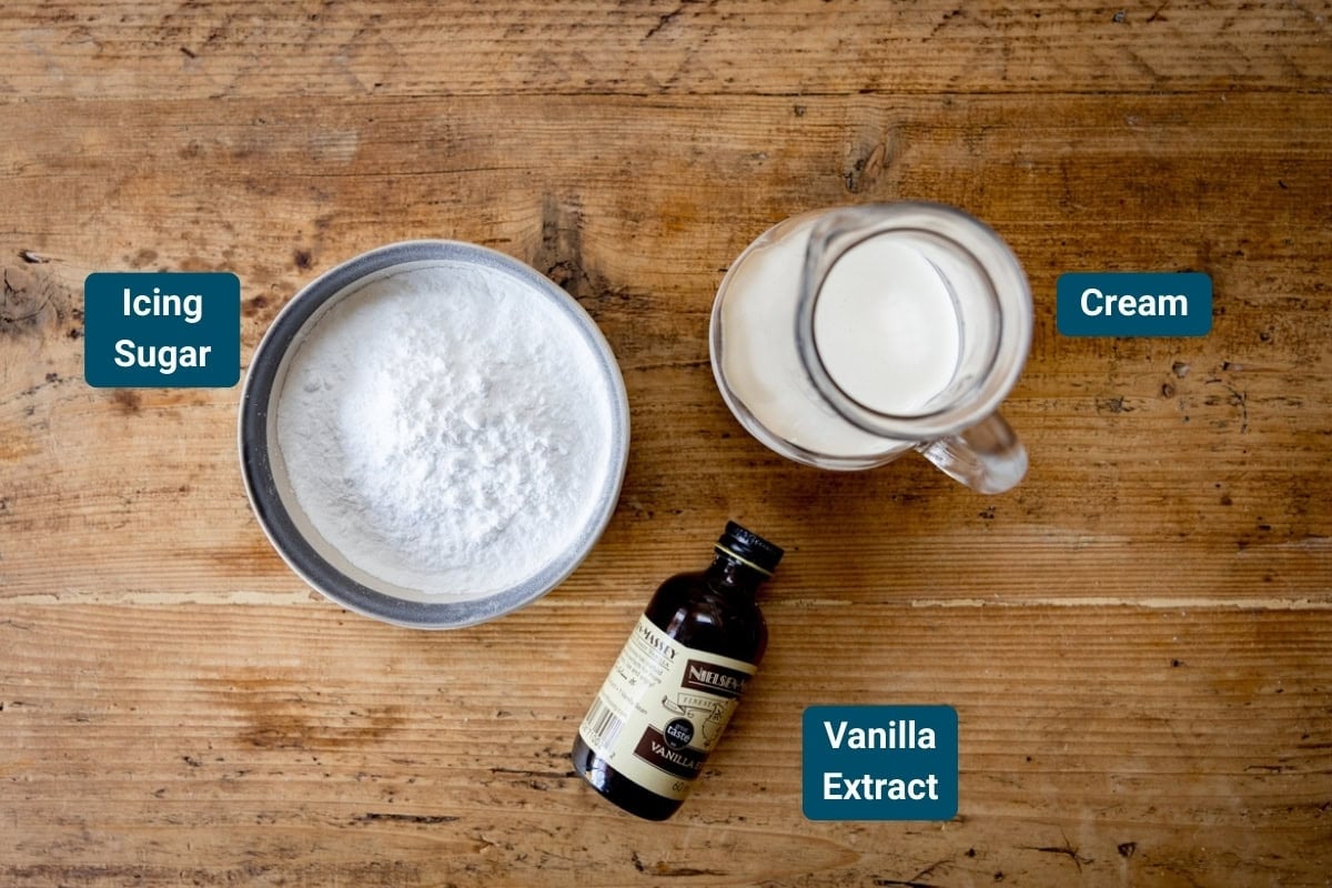 A bowl of icing sugar, a jug of cream, and a bottle of vanilla extract on a wooden table.