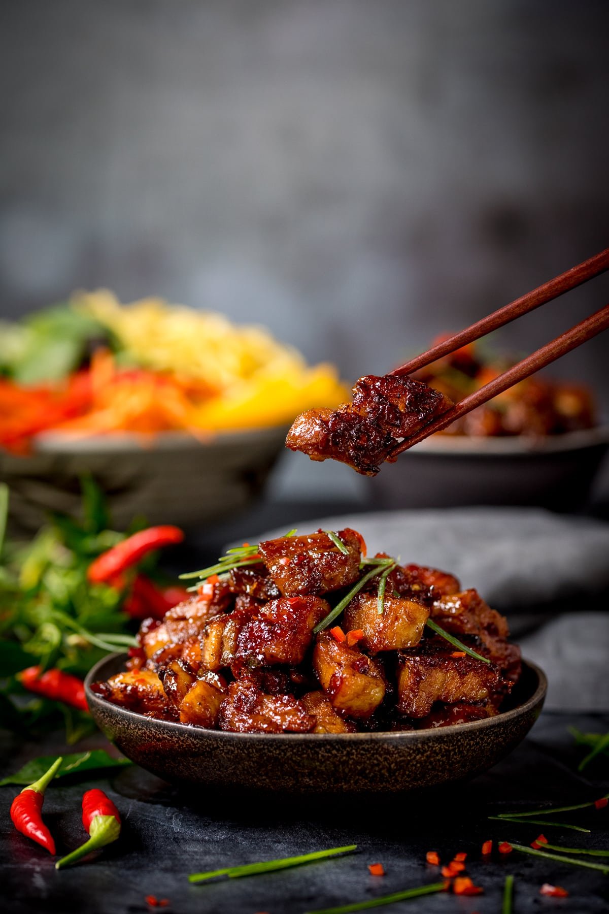 A bowl of Chinese sticky pork belly pieces against a dark background. There is a piece being lifted from the bowl with a pair of wooden chopsticks. There are ingredients scattered around and a bowl of colourful vegetables in the background.