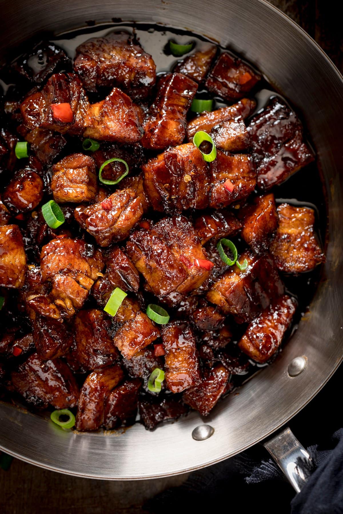 Close up overhead image of sticky Chinese pork belly, topped with chillies and spring onions in a silver frying pan.