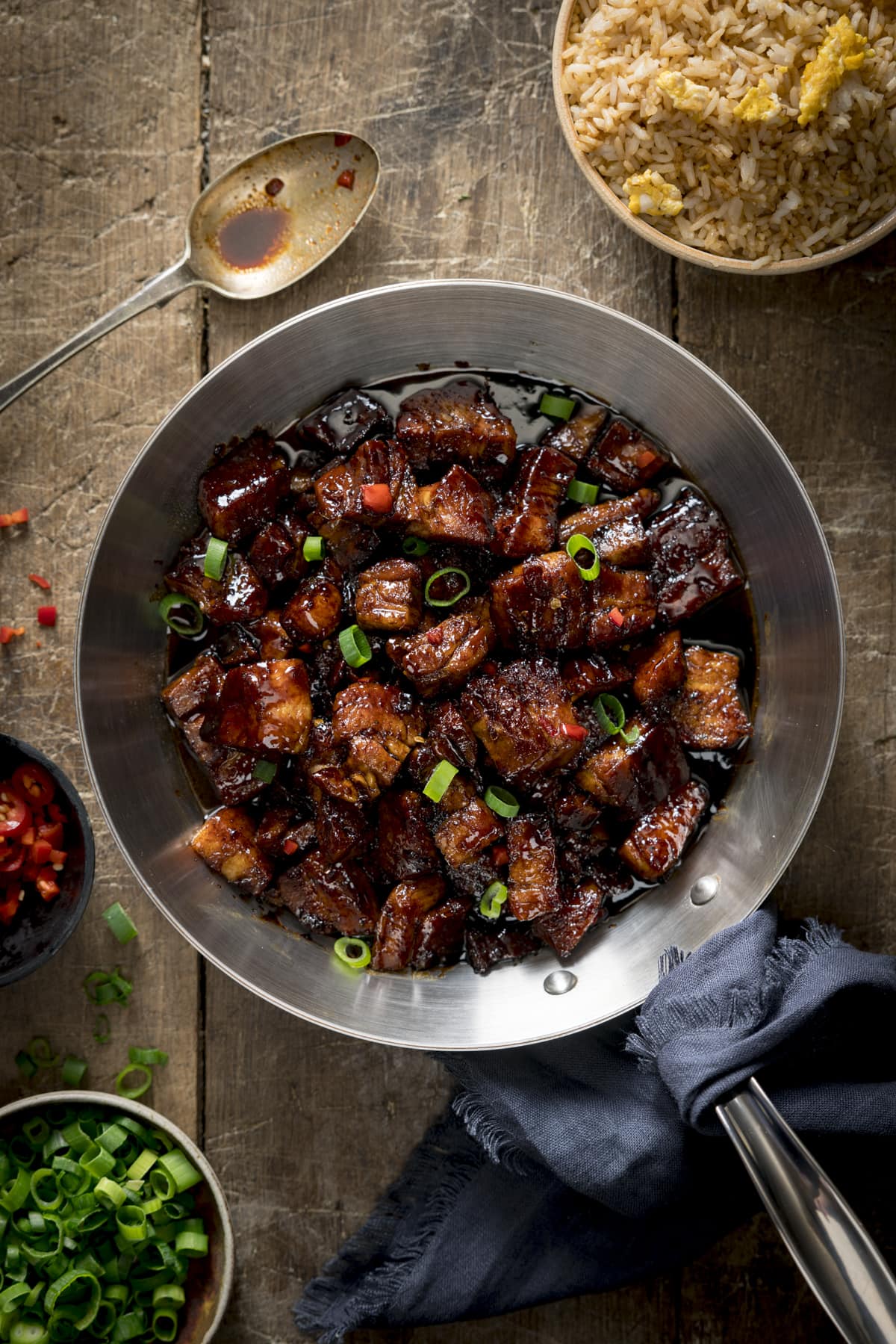 Overhead image of sticky Chinese pork belly in a silver frying pan. The pan is on a wooden table and there are spring onions, chillies and fried rice in bowls around the pan.