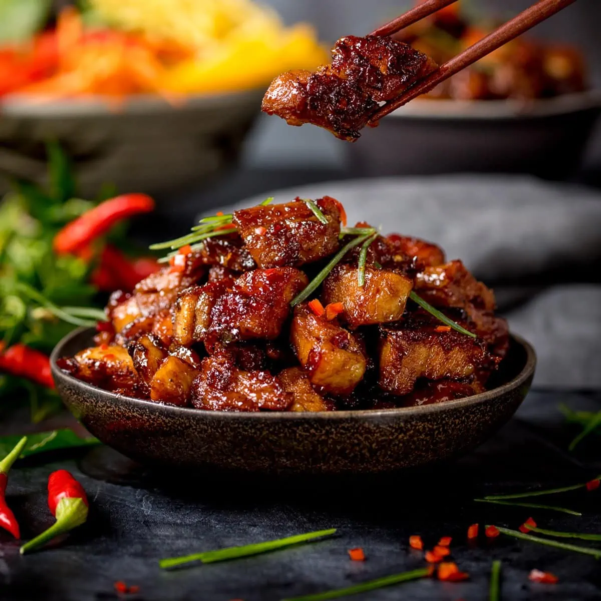 Close up image of bowl of Chinese sticky pork belly pieces. A piece is being taken from the bowl using a pair of chopsticks.