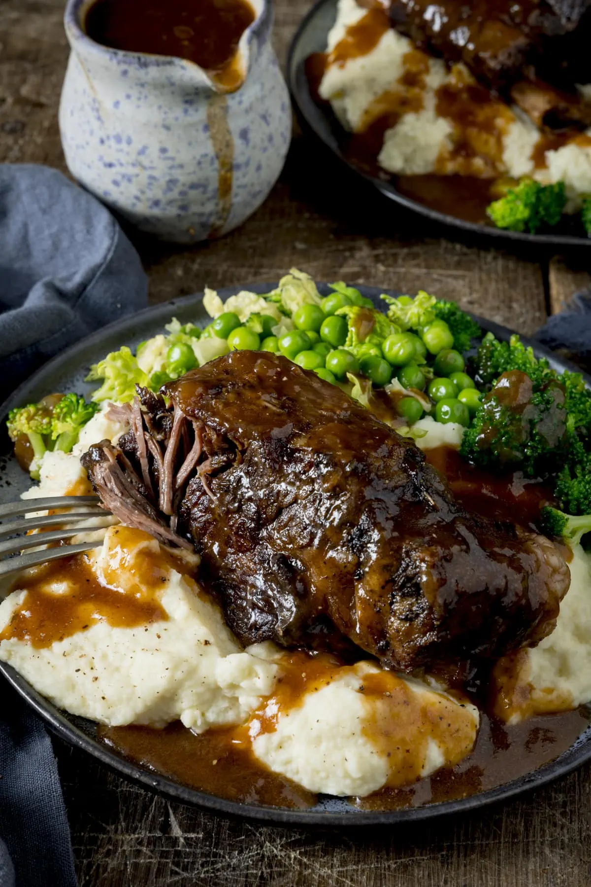 A close up of slow cooked beef short rib with gravy on a plate of mashed potatoes, with green vegetables also on the plate. The plate is on a wooden table. There is a forkful being taken from the short rib.
