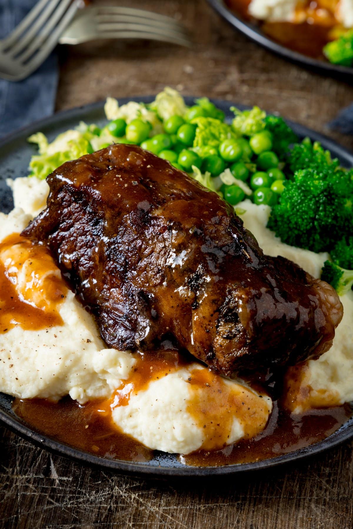A close up of slow cooked beef short rib with gravy on a plate of mashed potatoes, with green vegetables also on the plate. The plate is on a wooden table.