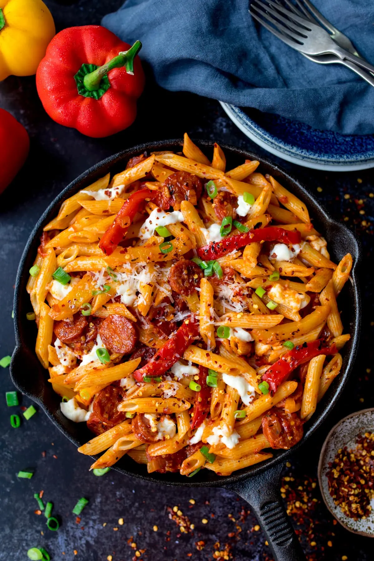 Overhead image of penne arrabbiata in a pan with mozzarella and chorizo. The pan is on a dark background and there are ingredients, plates, forks and a blue napkin scattered around.