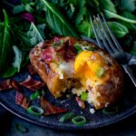 Square image of egg and bacon stuffed baked potato with runny egg on a dark plate