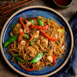 Overhead image of chicken lo mein on a blue plate on top of a wooden table. There is a pot of sweet chilli sauce and some chopsticks at the top of the image.