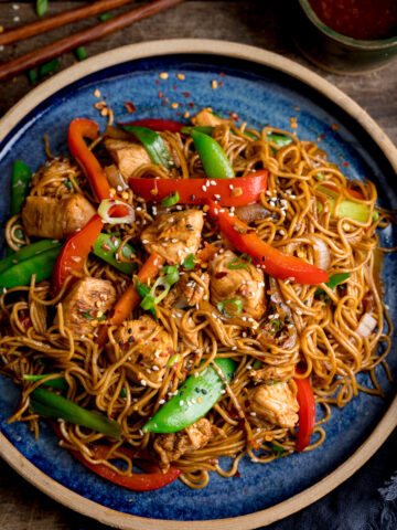 Overhead image of chicken lo mein on a blue plate on top of a wooden table. There is a pot of sweet chilli sauce and some chopsticks at the top of the image.