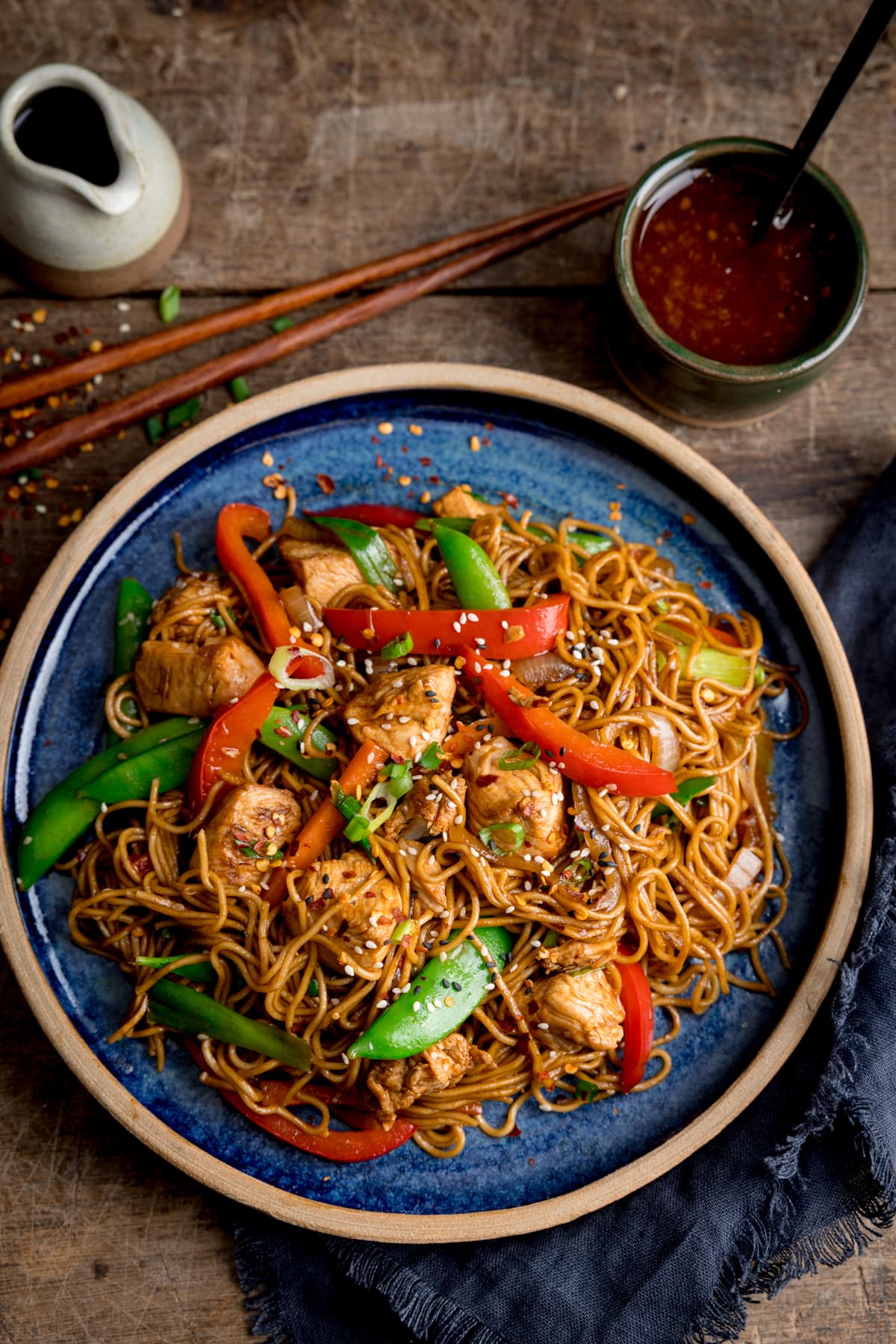 Overhead image of Chicken Lo Mein on a blue plate on a wooden table.  At the top of the picture are a pot of sweet chili sauce, a small jar of soy sauce, and some chopsticks.