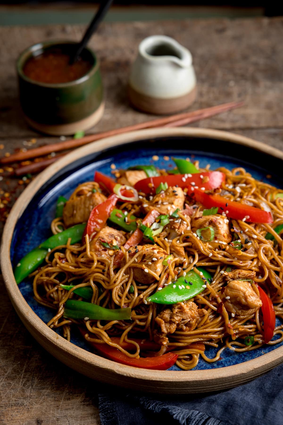 Side image of chicken lo mein on a blue plate on top of a wooden table. There is a pot of sweet chilli sauce, with a spoon sticking out, a little jug of soy sauce and some chopsticks at the top of the image.
