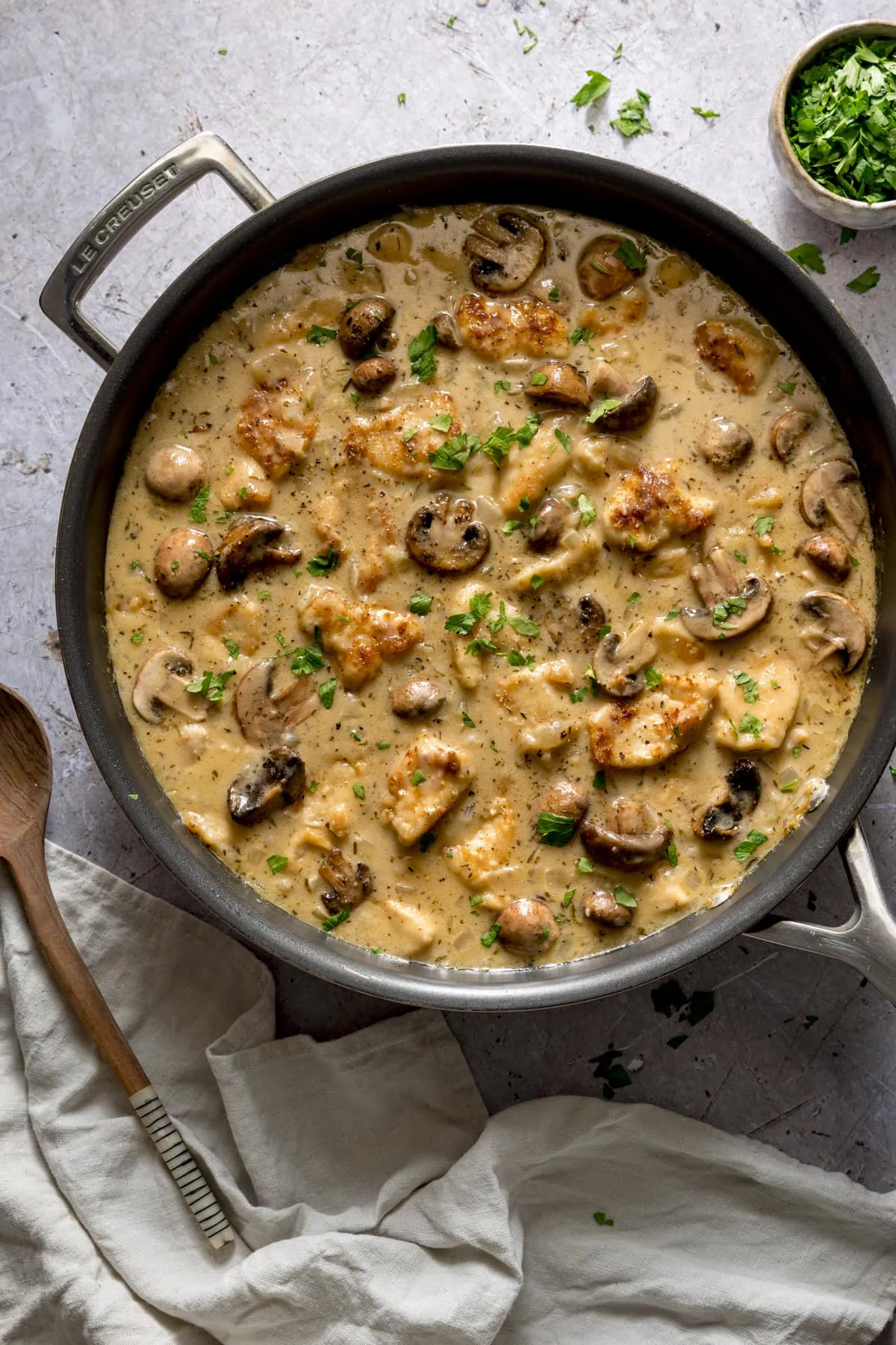 Overhead image of creamy chicken and mushroom casserole in a large frying pan on a light background. There is a cream linen napkin and a wooden spoon next to the pan and parsley scattered in and around the pan.