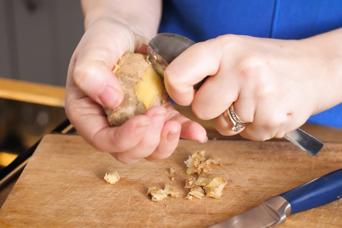 A person uses a spoon to peel fresh ginger over a wooden chopping board, preparing ingredients for Ginger Beef Stir-Fry, with ginger peelings and a knife nearby.